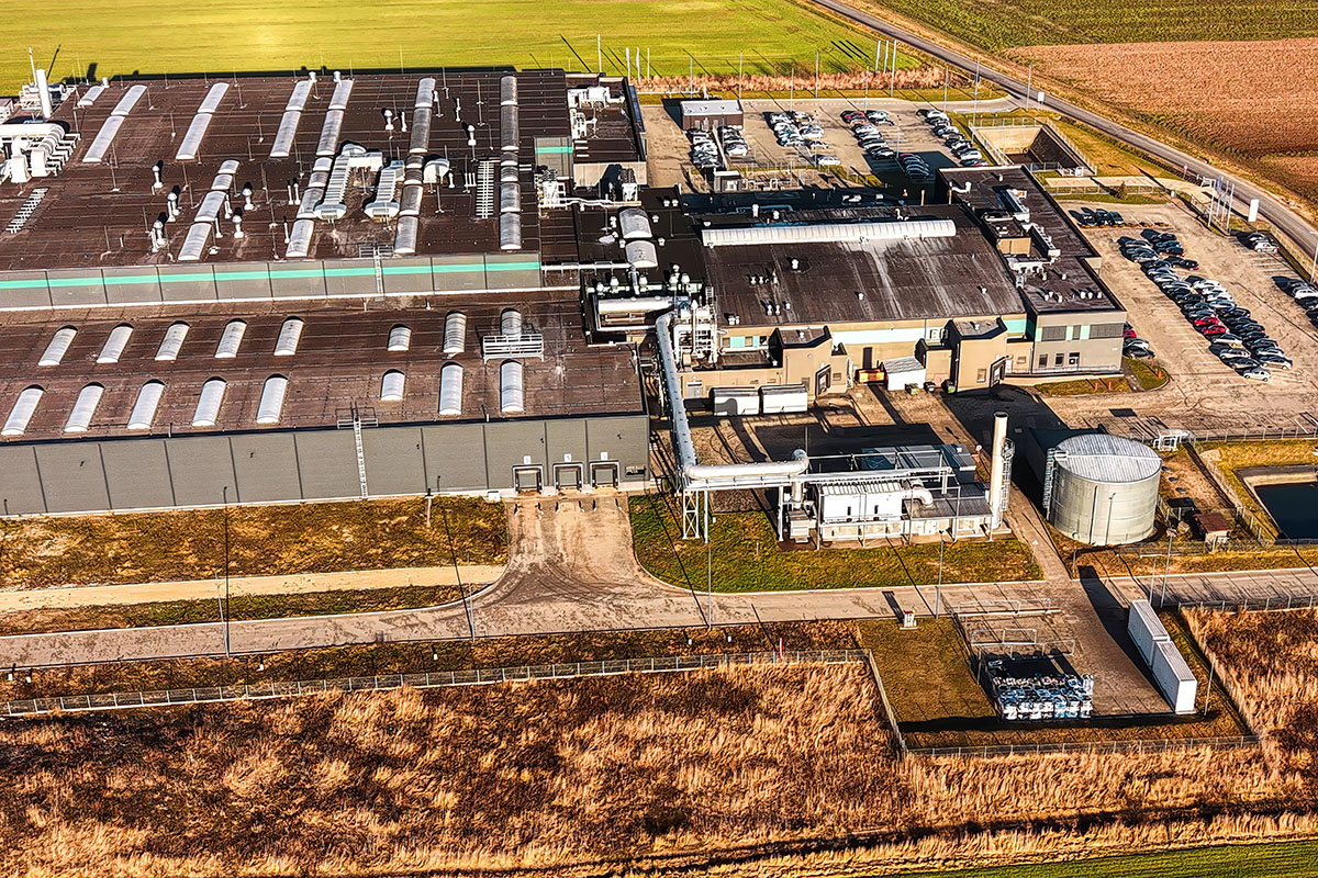 Aerial view of a large industrial facility with rooftop ventilation units, a cylindrical storage tank and a car park, surrounded by open farmland and dry grassland