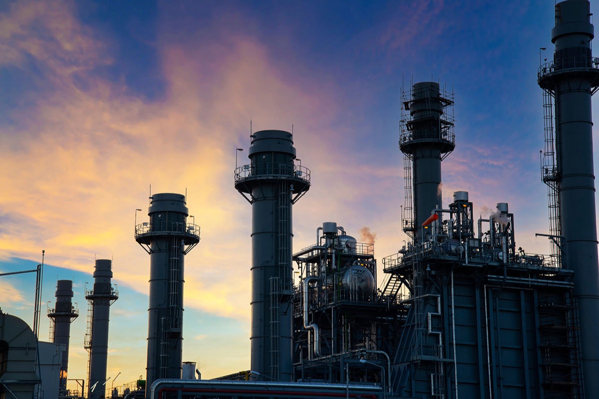 Tall exhaust stacks and steel structures of a gas-fired power plant silhouetted against a blue and orange sky at dusk