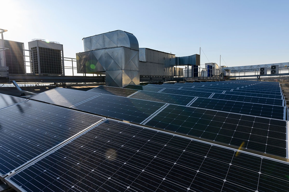 Rows of solar panels on a flat commercial roof beside large metal ventilation ducts and rooftop air conditioning units under a clear sky