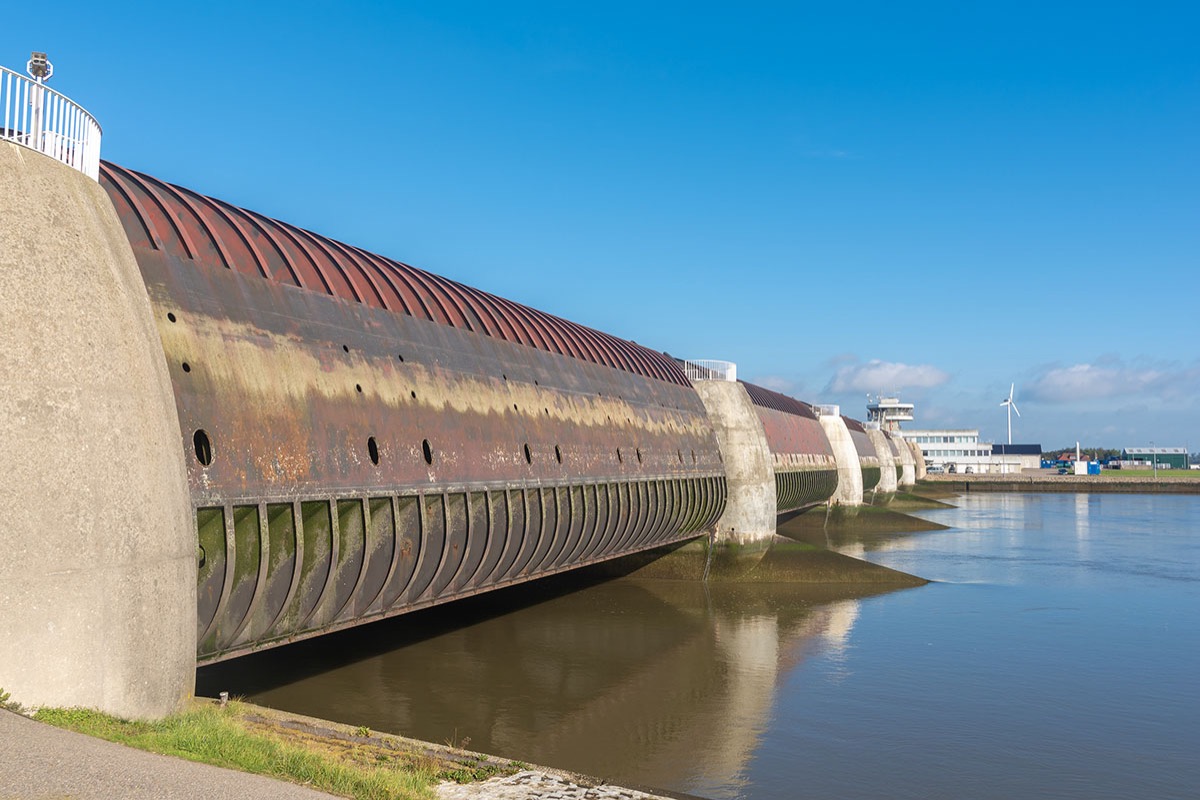 A steel and concrete storm surge barrier with curved sluice gates stretching across a tidal estuary under a clear blue sky