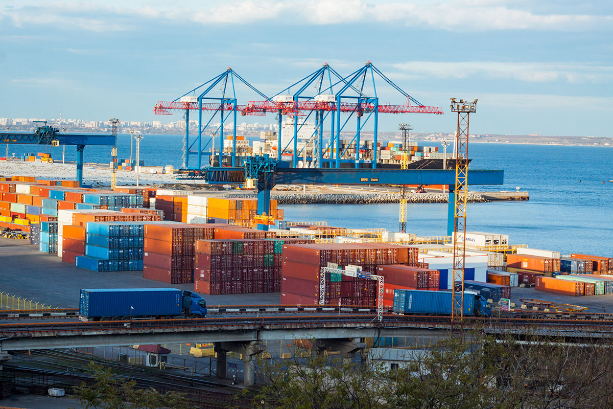 Container cranes and stacked shipping containers at a busy seaport, with trucks moving along the terminal road
