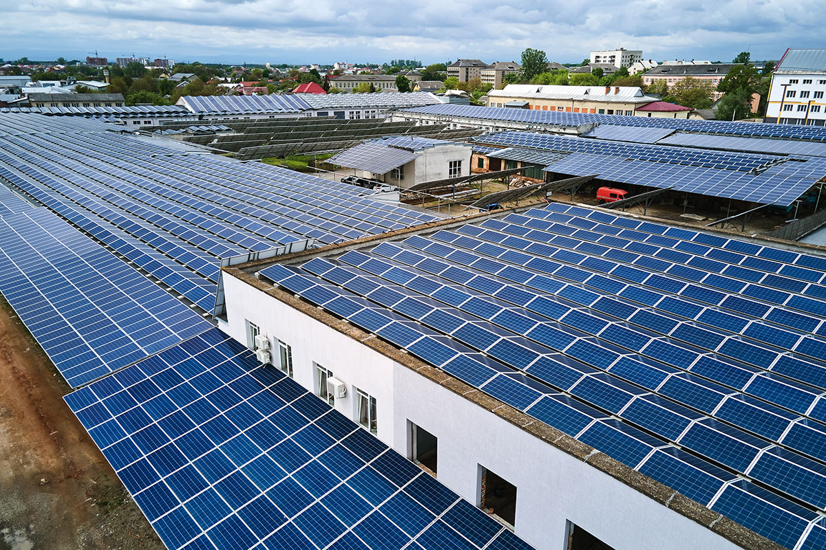 Aerial view of rows of blue solar panels covering the flat roofs of several industrial buildings on the edge of a town