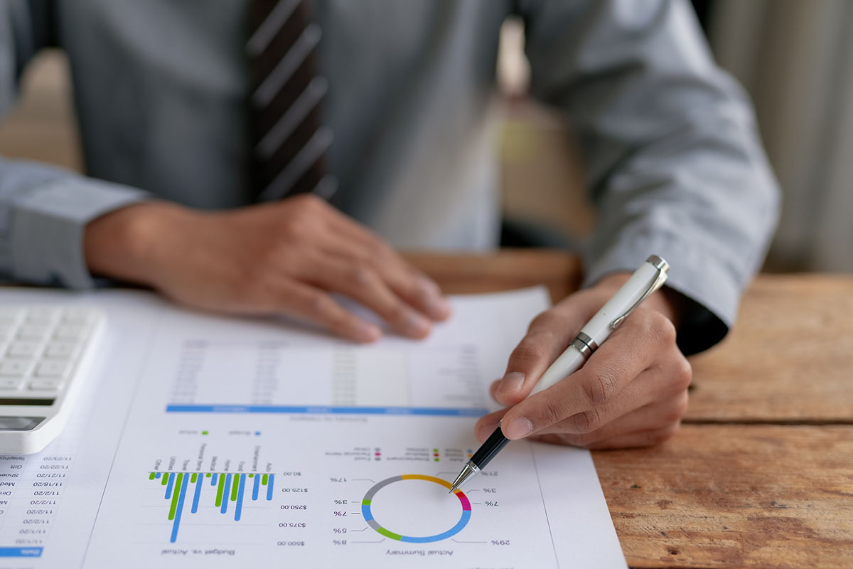 Close-up of a businessman in a shirt and tie pointing a white pen at a printed report of bar charts and a ring chart on a wooden desk beside a calculator
