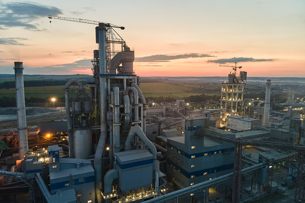 Aerial view of a cement plant at dusk with a tall preheater tower, ducting, smokestacks and two construction cranes against a pink and orange sky over farmland