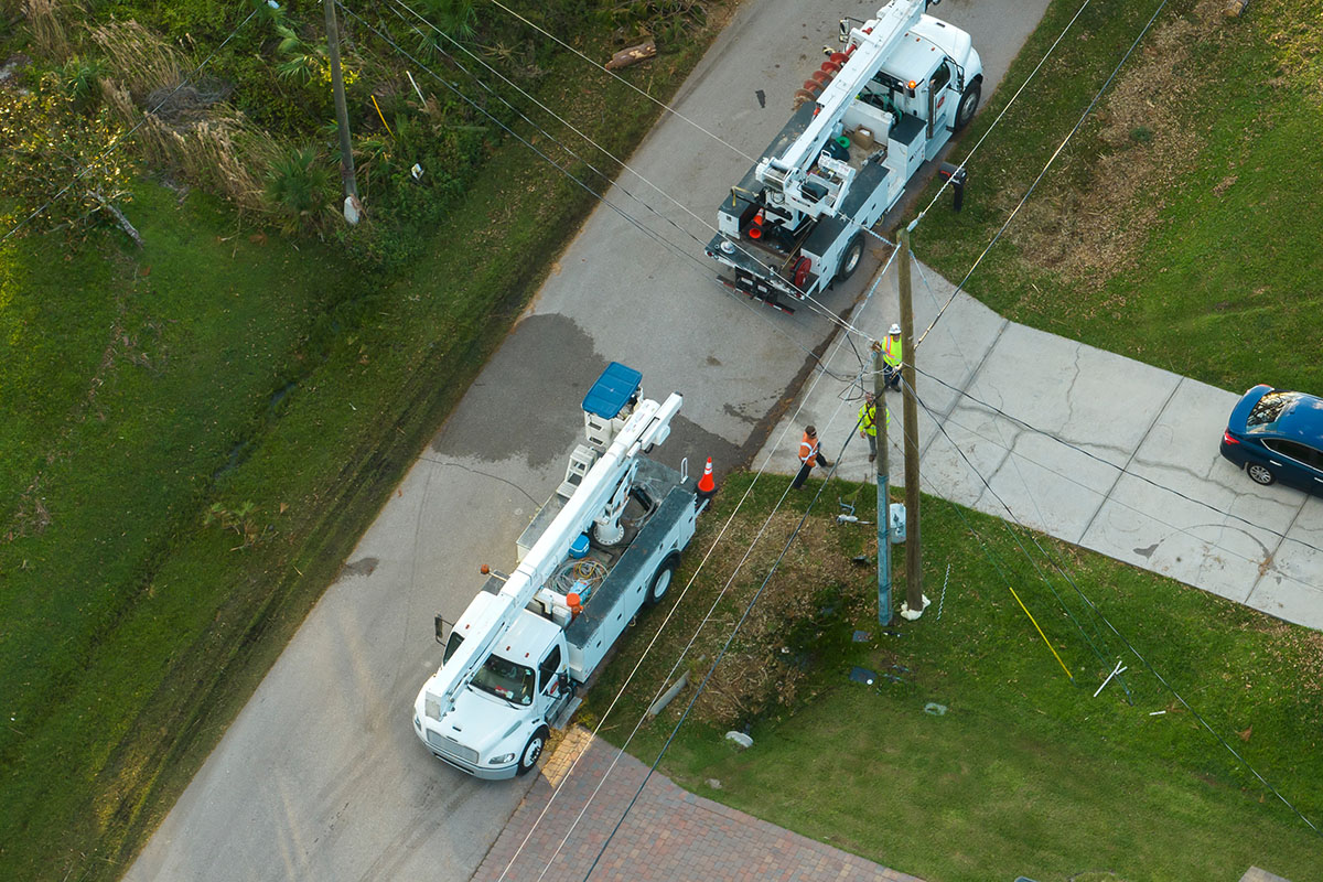 Aerial view of two utility bucket trucks and a line crew restoring power at a roadside pole in a Florida neighborhood after a hurricane