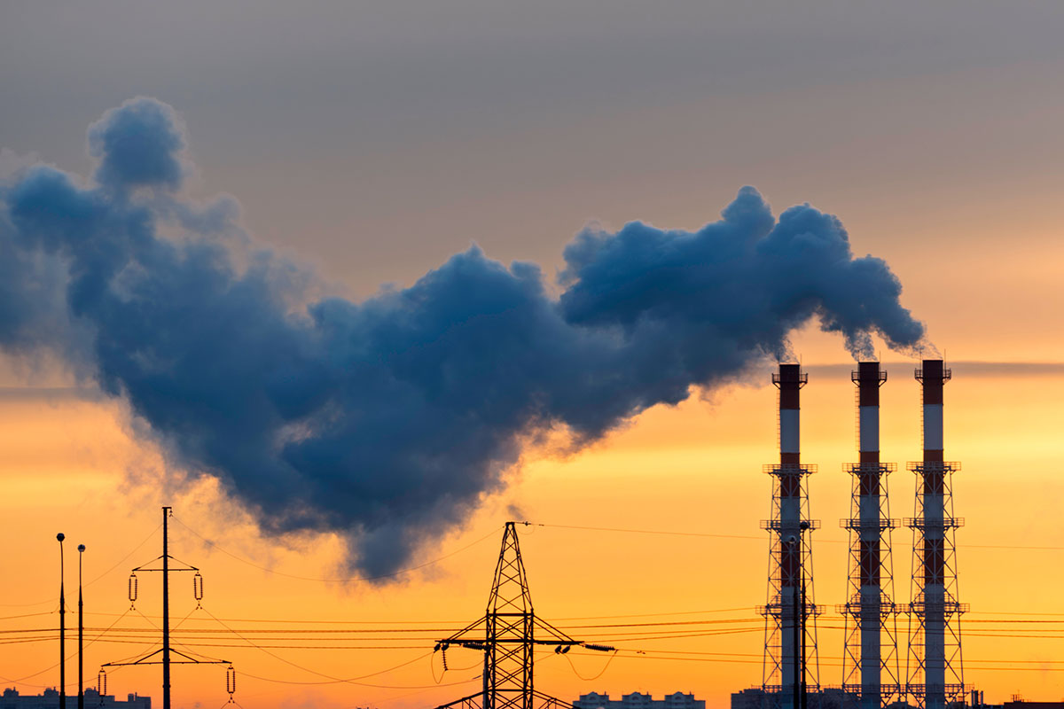 A large plume of dark smoke drifting from three red and white power plant stacks across an orange sunset sky, with transmission towers and power lines below